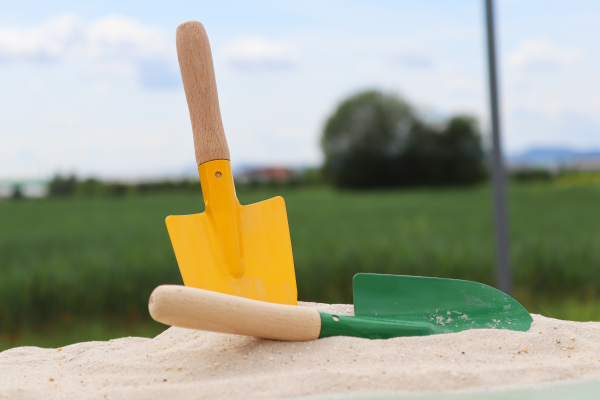 Zwei Sandschaufeln für Kinder stecken  im Sand eines Spielplatzes. Sie haben einen gelben und grünen Besatz. Dahinter befindet sich eine Wiese.